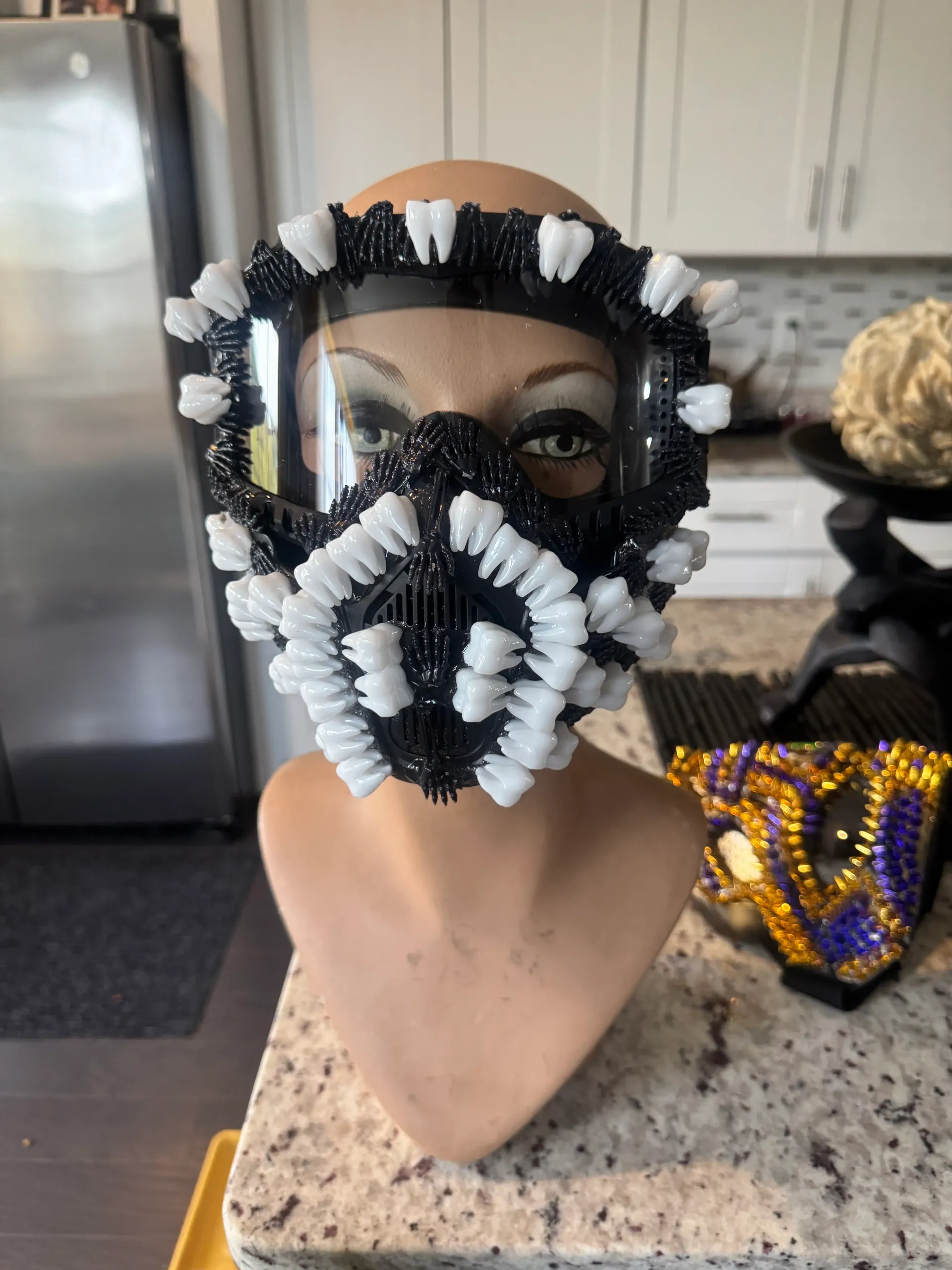 Front view of the black spike-armored mask with rows of white teeth and a clear visor on a mannequin bust