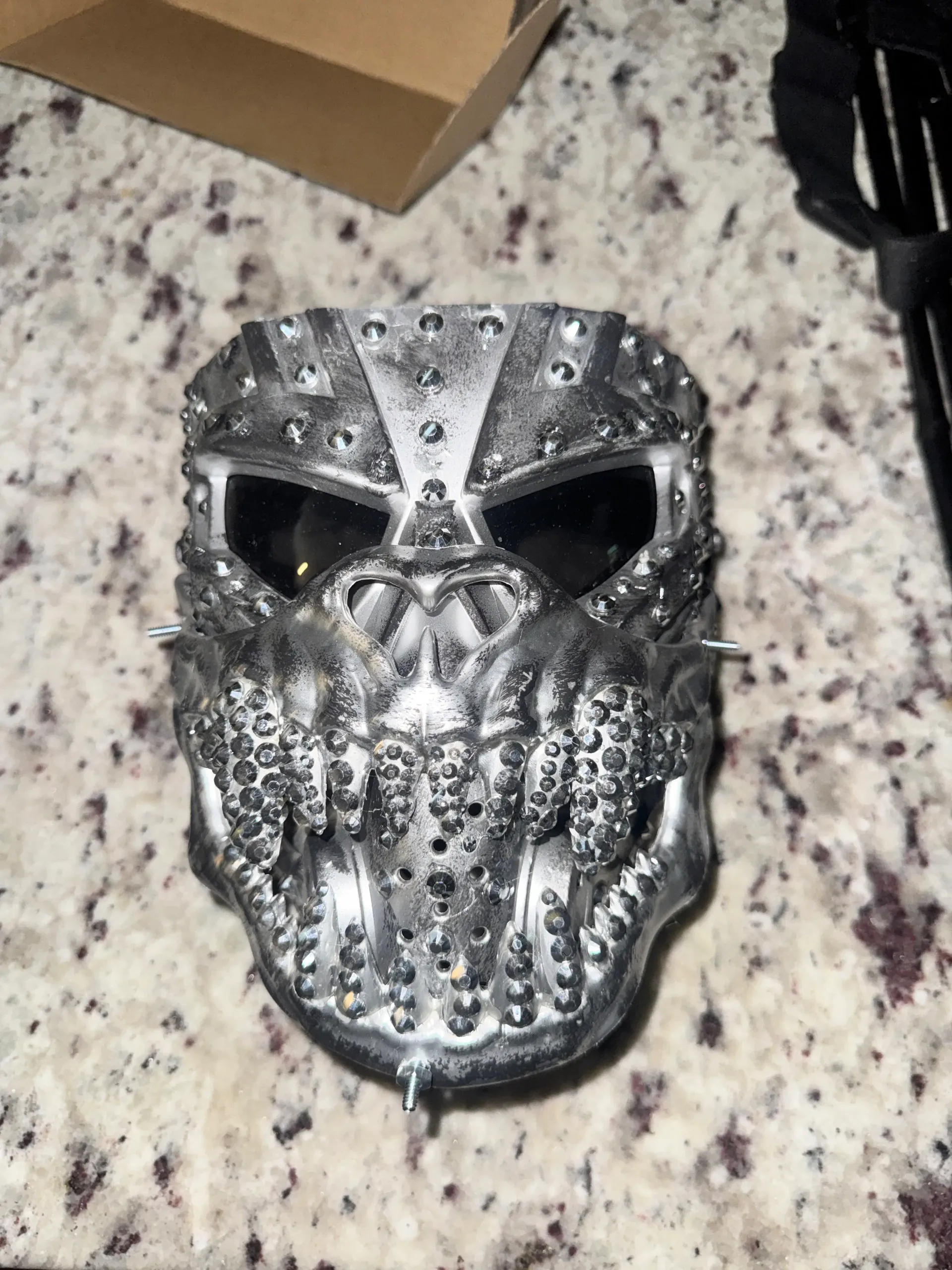 Close-up detail of the silver two-face mask with dark lenses, layered metal plating, studs, and crystal accents